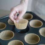 Hand placing brown cupcake liner in muffin baking tray, ready for baking.