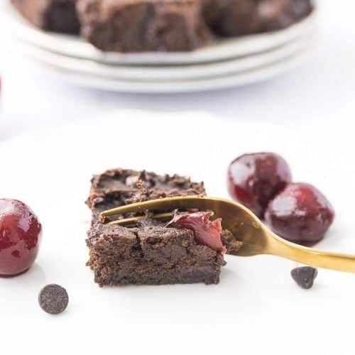 a gold fork taking a piece of cherry chocolate protein brownie on a white plate. Stack of brownies in background.