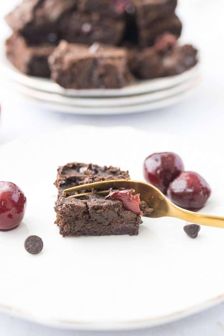 a gold fork taking a piece of cherry chocolate protein brownie on a white plate. Stack of brownies in background.