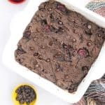 baked cherry chocolate protein brownies in pan, uncut. small bowl of chocolate chips and small bowl of cherries for decoration in the photo.