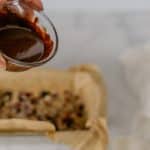 hand pouring melted chocolate over kind style energy bars dry ingredients in parchment lined loaf pan