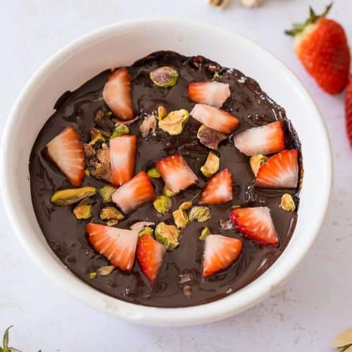 Dubai Chocolate Protein Brownie Mug Cake close up in rameking with chopped strawberries and pistachios on top and scattered around dish