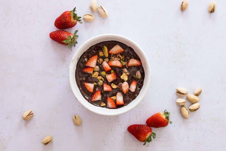 Image of Dubai Chocolate Protein Brownie Mug Cake in white ramekin topped with strawberries and strawberries and shelled pistachios scattered around