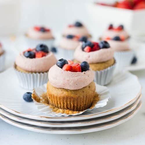 refined sugar free strawberry shortcake cupcake topped with cashew frosting and blueberries and strawberries on 3 white plates with gold edge with white wrapper unwrapped exposing the cupcake.