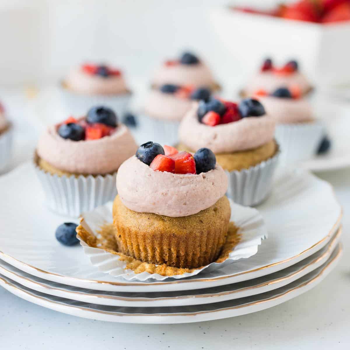 refined sugar free strawberry shortcake cupcake topped with cashew frosting and blueberries and strawberries on 3 white plates with gold edge with white wrapper unwrapped exposing the cupcake.
