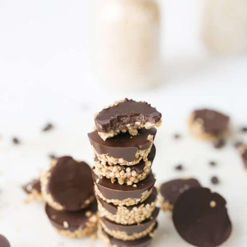 6 SunButter Oat cups stacked with others scattered around. Mason jars with oats and quinoa in the background.