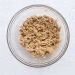 Puffed quinoa, oats, sunflower butter, maple syrup, cinnamon, and salt mixed together in large clear mixing bowl on white background.