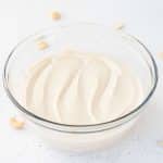Vegan Cashew Frosting in clear glass bowl with cashews scattered around on white surface.