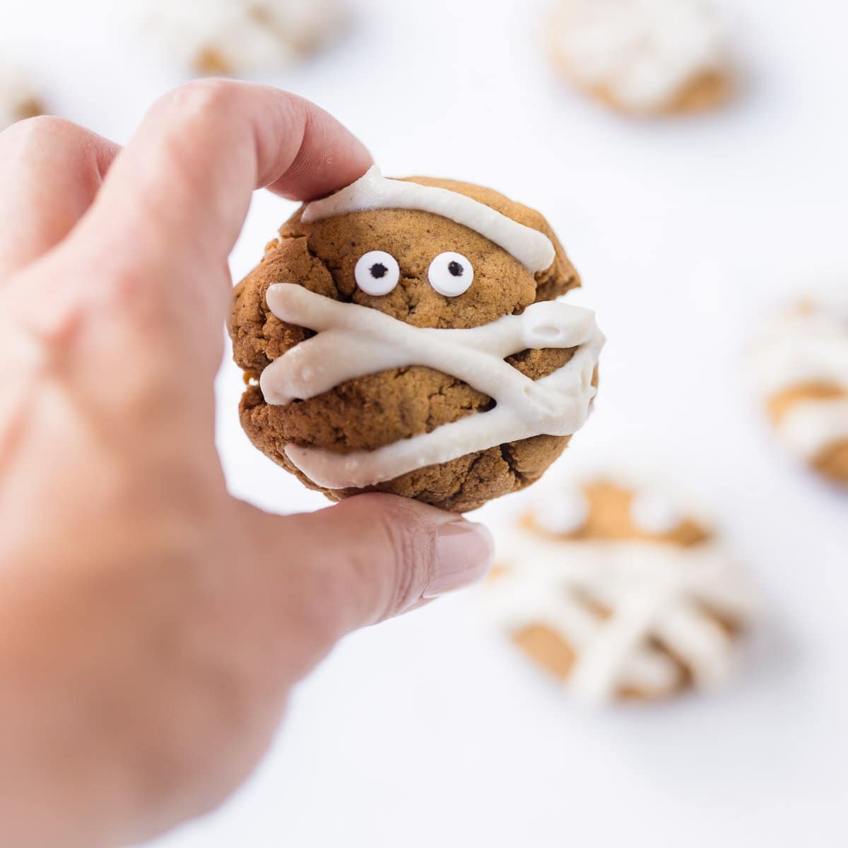 hand holding up a single vegan pumpkin pie spice cookie with other cookies softly blurred in background on white surface