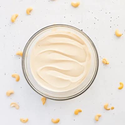 Vegan Cashew Frosting in clear glass bowl with cashews scattered around on white surface.