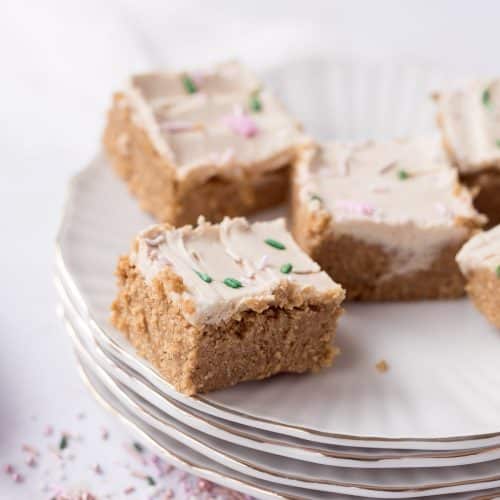 Five soft, sugar cookie protein bars stacked on white vintage plates with gold rims, topped with creamy protein frosting and festive sprinkles.