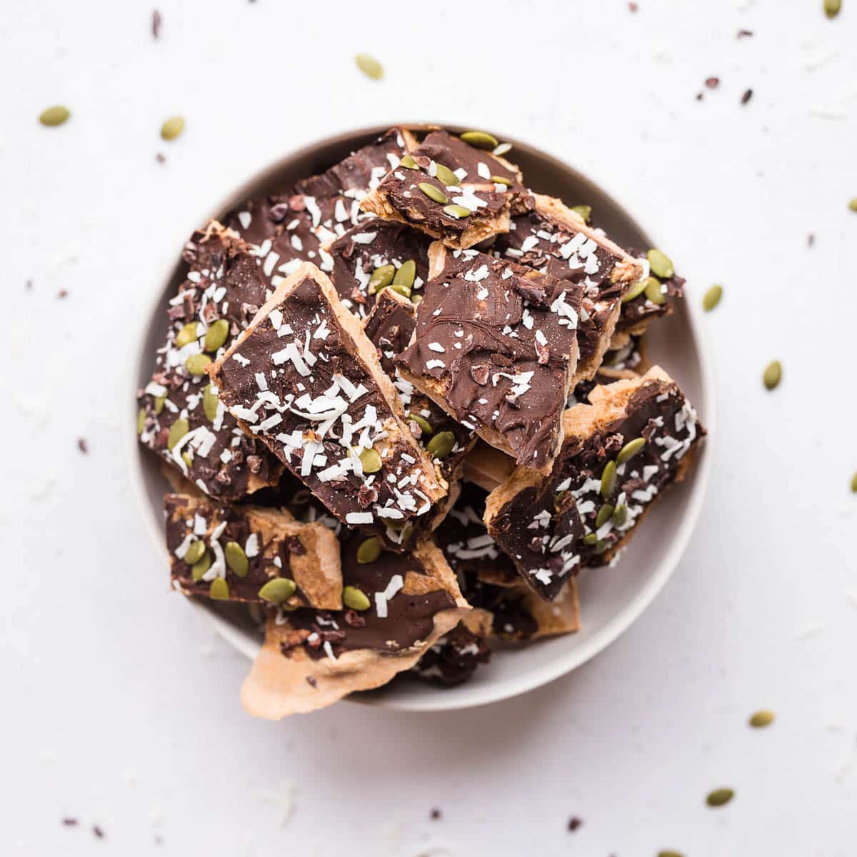 pumpkin spice banana yogurt bark in bowl on white surface with cacao nibs and pumpkin seeds around