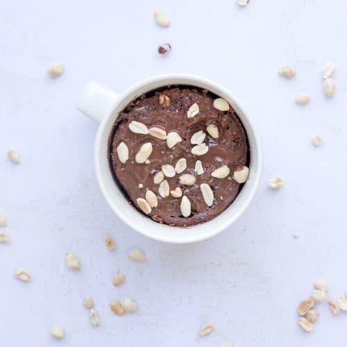 High protein Snickers mug cake topped with melted chocolate, crushed peanuts, and creamy caramel for a quick healthy dessert.
