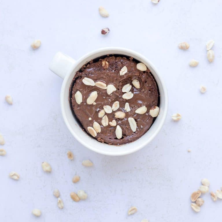High protein Snickers mug cake topped with melted chocolate, crushed peanuts, and creamy caramel for a quick healthy dessert.