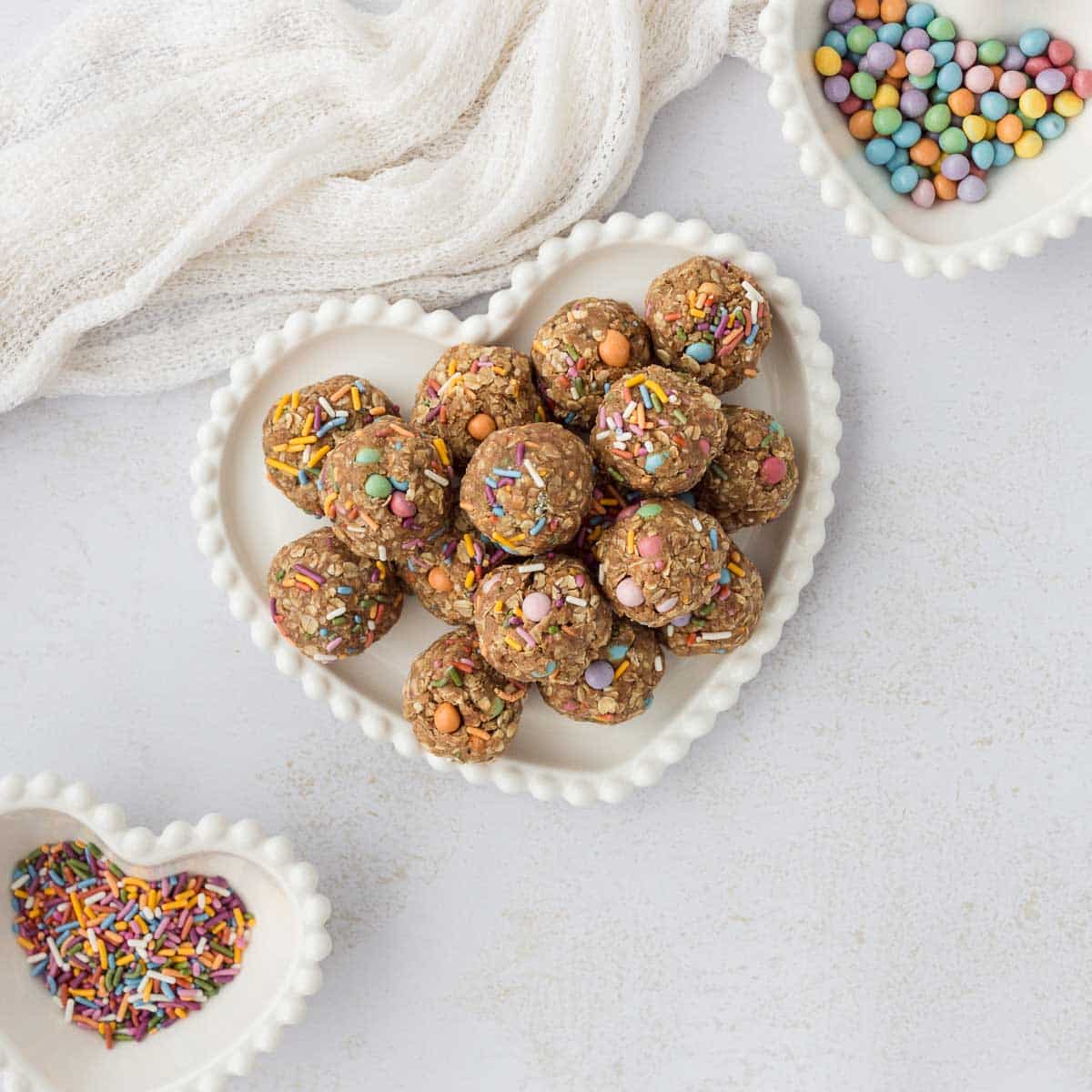 birthday cake funfetti protein balls on a decorative white heart plate with 2 small white heart bowls filled with sprinkled and candy chips
