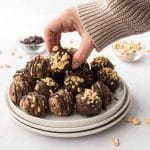 Hand taking a chocolate-coated Snickers protein ball from white plate after having chilled in fridge