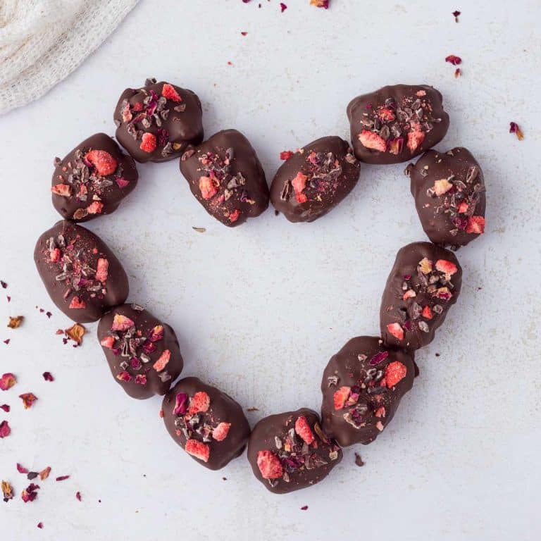 Strawberry Cheesecake Chocolate Covered Stuffed Dates shaped into a heart on white surface with pieces of rose petals around