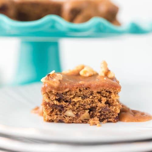Date Cake with Caramel Sauce slice on white plates with whole cake on cake stand blurred in background