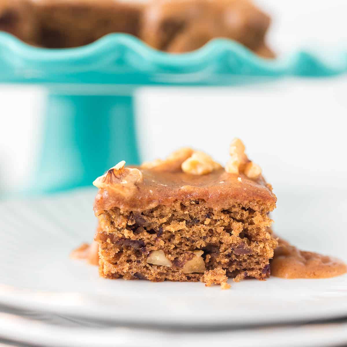 Date Cake with Caramel Sauce slice on white plates with whole cake on cake stand blurred in background