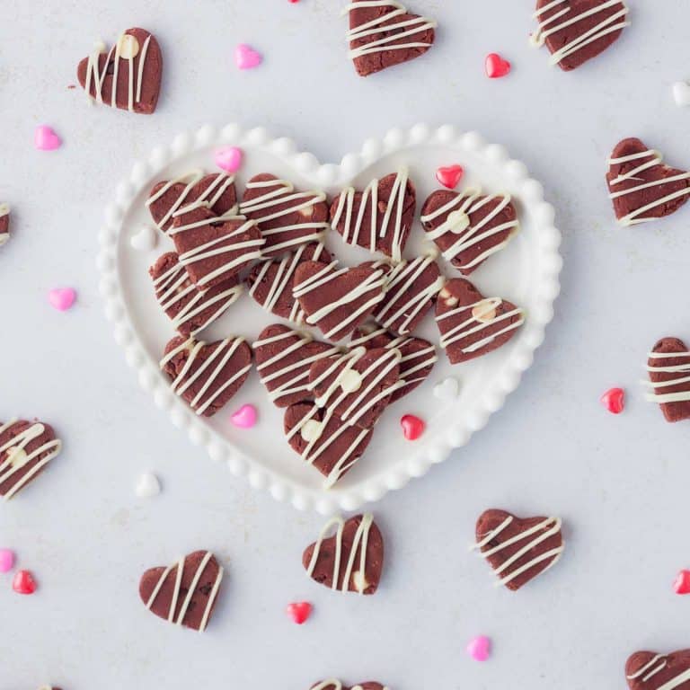 red velvet cookie dough hearts on white decorative heart plate with edible cookie dough hearts and heart sprinkles around on white surface