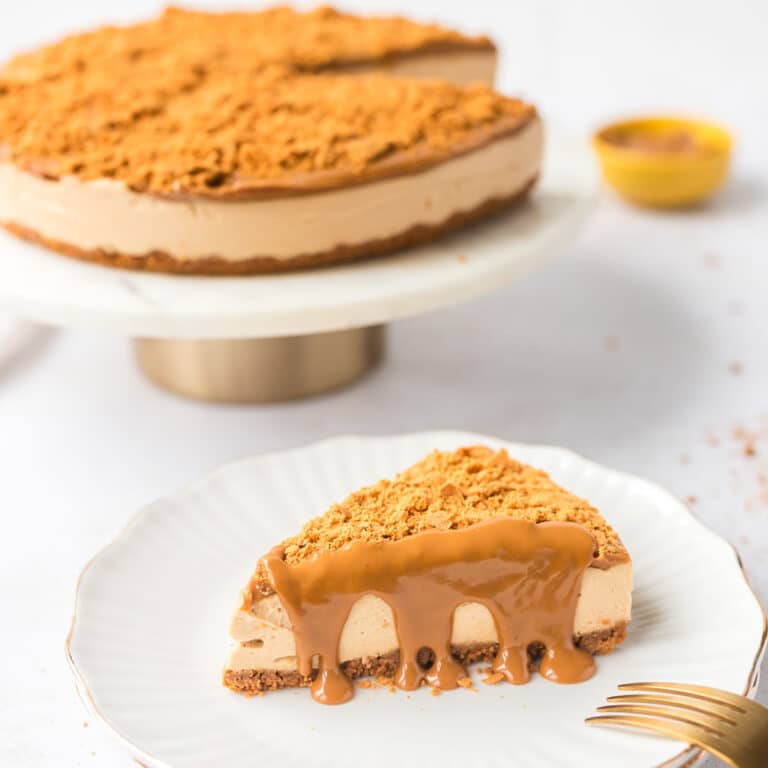 Slice of High Protein No Bake Biscoff Cheesecake on white plate with spread dripping down and rest of cheesecake in background on cake stand